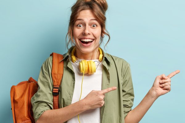 Horizontal shot of glad Caucasian woman points aside with both fore fingers, advertises something aside, dresed in stylish clothes, carries rucksack, listens music in modern headphones. Promotion